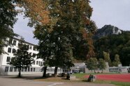 Vor dem Schulgebäude stehen Bäume. Der Sportplatz ist mit auf dem Gelände.