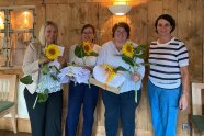 Drei Frauen mit Sommenblumen und eine Frau ohne Sonnenblume stehen lachend vor einer Holzwand.