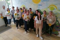 Vor einem bunten Bild mit Sonnenschein stehen die Teilnehmer und halten eine Sonnenblume in der Hand